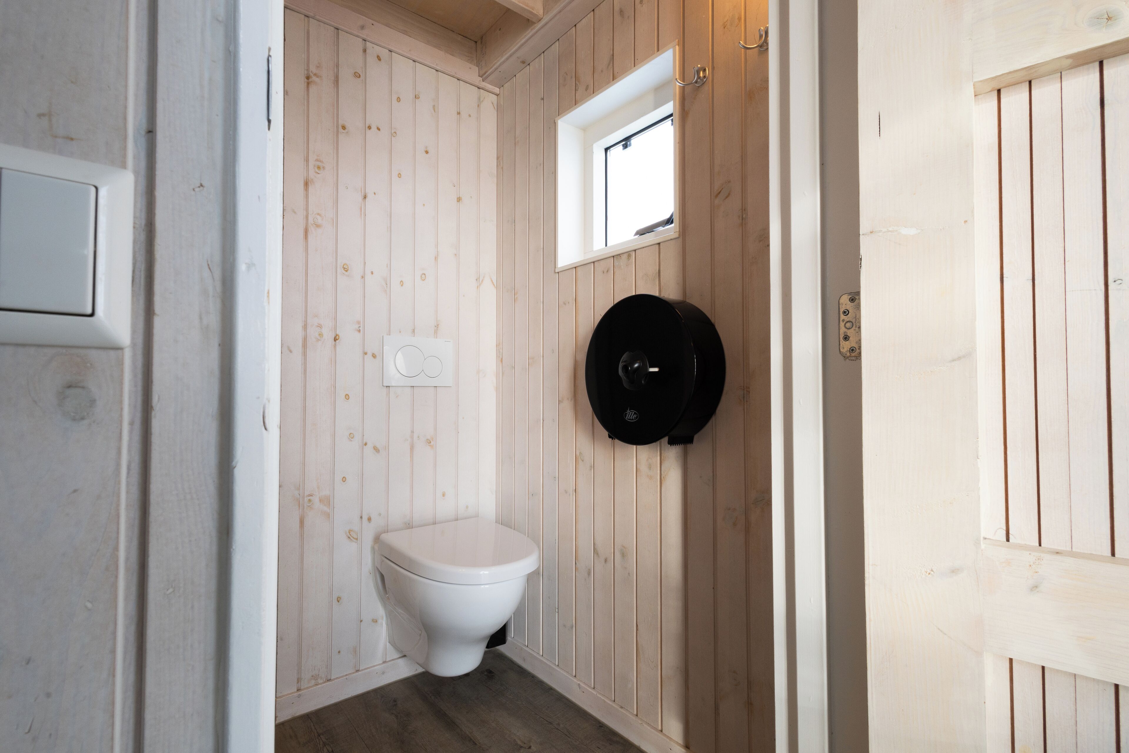 Une petite salle de bain compacte avec des murs en bois, des toilettes blanches, une fenêtre et un objet circulaire noir accroché au mur.