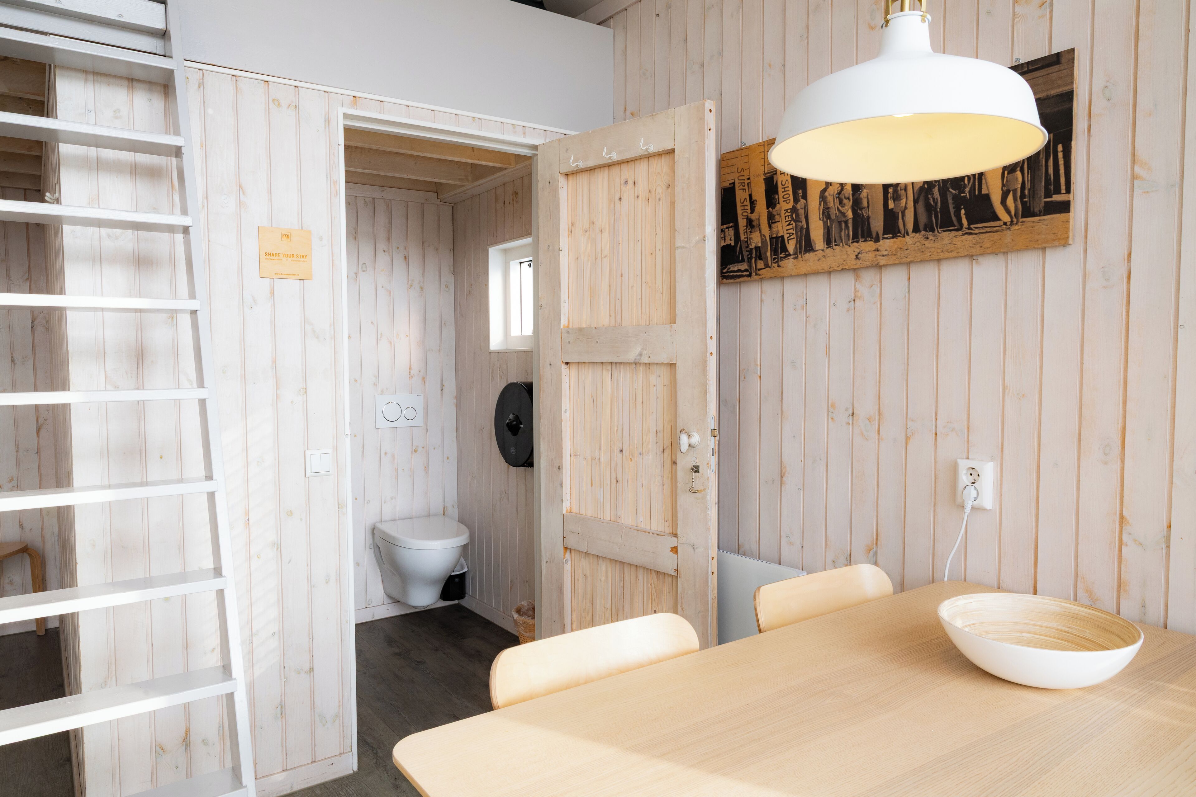 A room with a wooden interior, a white table with a bowl, a white chair, a white ceiling lamp, and a wooden door leading to another room.