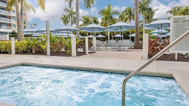 Una piscina con escalera metálica, rodeada de palmeras y sombrillas.