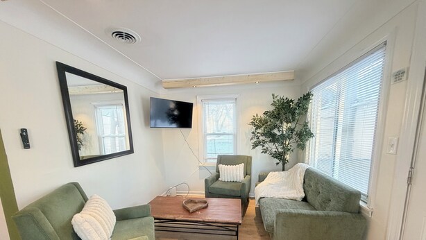 Una sala de estar con dos sillones verdes, una mesa de centro de madera, un televisor montado en la pared y un ventanal con persianas.