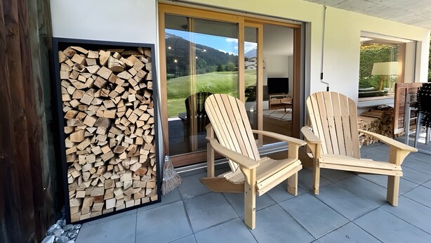 Eine überdachte Terrasse mit zwei Holz-Adirondack-Stühlen, einem Holzscheitestapel und einer Schiebetür zu einem Außenbereich.