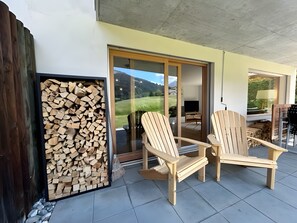 Eine überdachte Terrasse mit zwei Holz-Adirondack-Stühlen, einem Holzscheitestapel und einer Schiebetür zu einem Außenbereich.