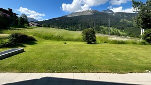 Ein grasbewachsenes Feld mit einer Straße, einem Berg im Hintergrund und einem Gebäude mit Satteldach.