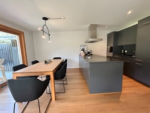 Eine moderne Küche mit einer zentralen Kochinsel, dunklen Schränken und einem Essbereich mit Holztisch und schwarzen Stühlen.