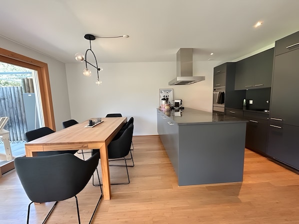 Eine moderne Küche mit einer zentralen Kochinsel, dunklen Schränken und einem Essbereich mit Holztisch und schwarzen Stühlen.