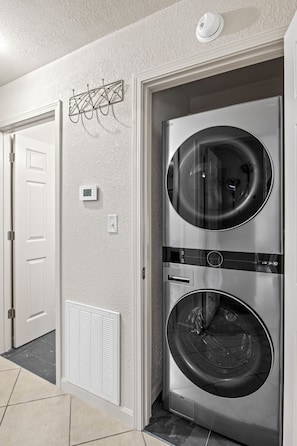 A laundry room with two stacked front-loading washing machines, a white door, a wall-mounted thermostat, and a ceiling-mounted smoke detector.