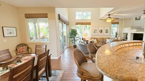 A spacious kitchen with a curved granite countertop, a dining area with wicker chairs, and a view of the outdoors through large windows. - Hilton Waikoloa Pool Pass 2026-27, Beach and Baby Gear! (Waikoloa Village)