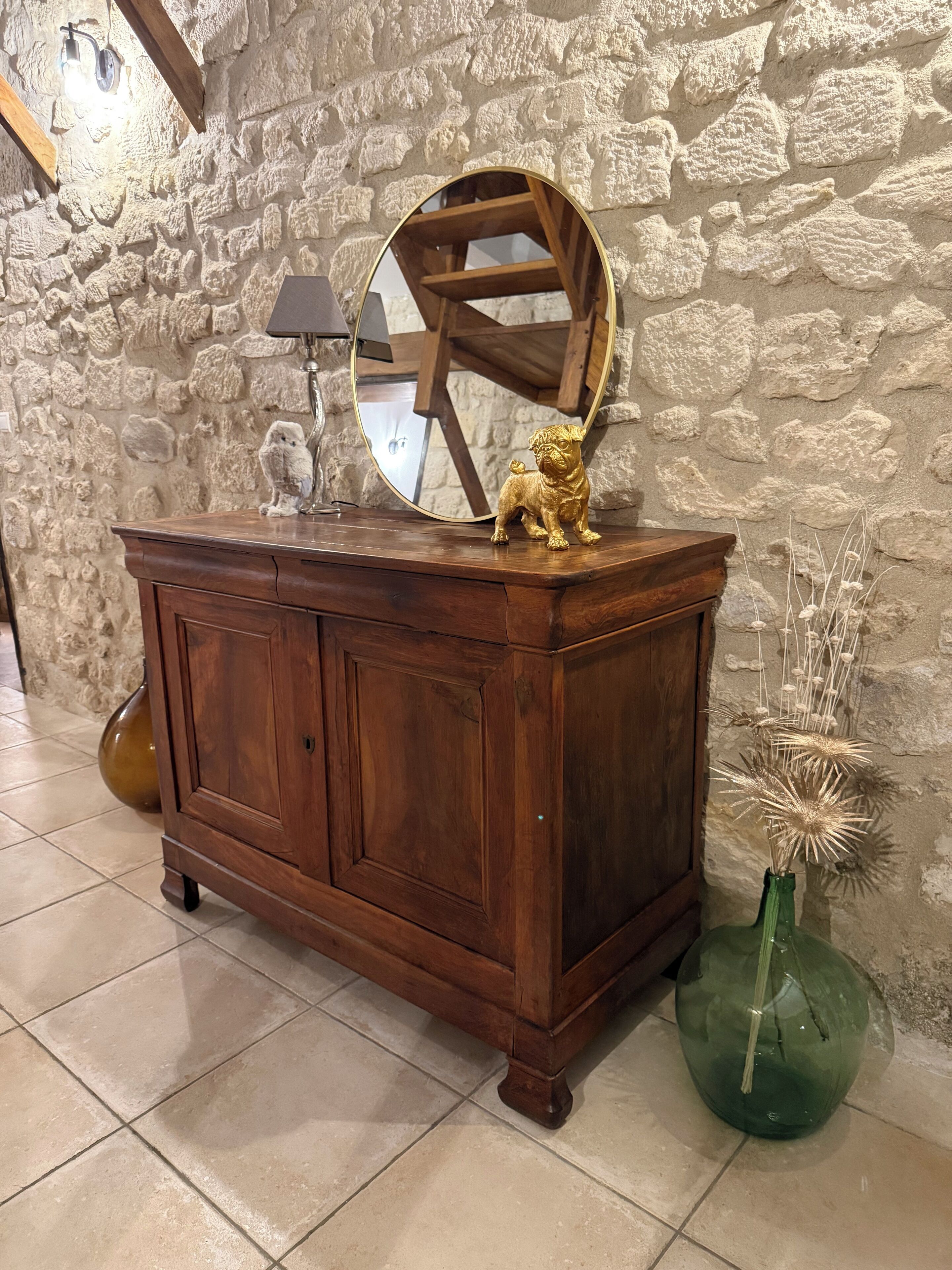 Un mueble de madera con un espejo, una pequeña figura de perro dorado y un jarrón verde con plantas secas.