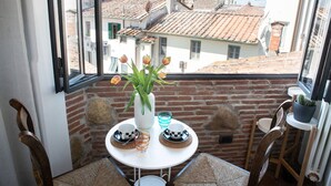 Ein Eckbalkon mit einem weißen Tisch für zwei Personen, einer Blumenvase und Blick auf Häuser mit Terrakotta-Dächern.