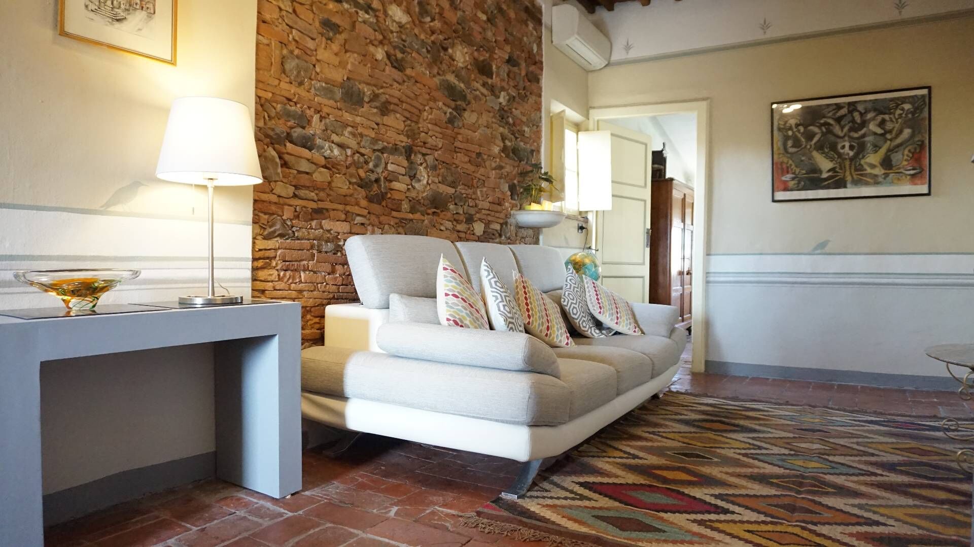 A living room with a stone wall, a sofa with patterned cushions, a side table with a lamp, and a patterned rug.