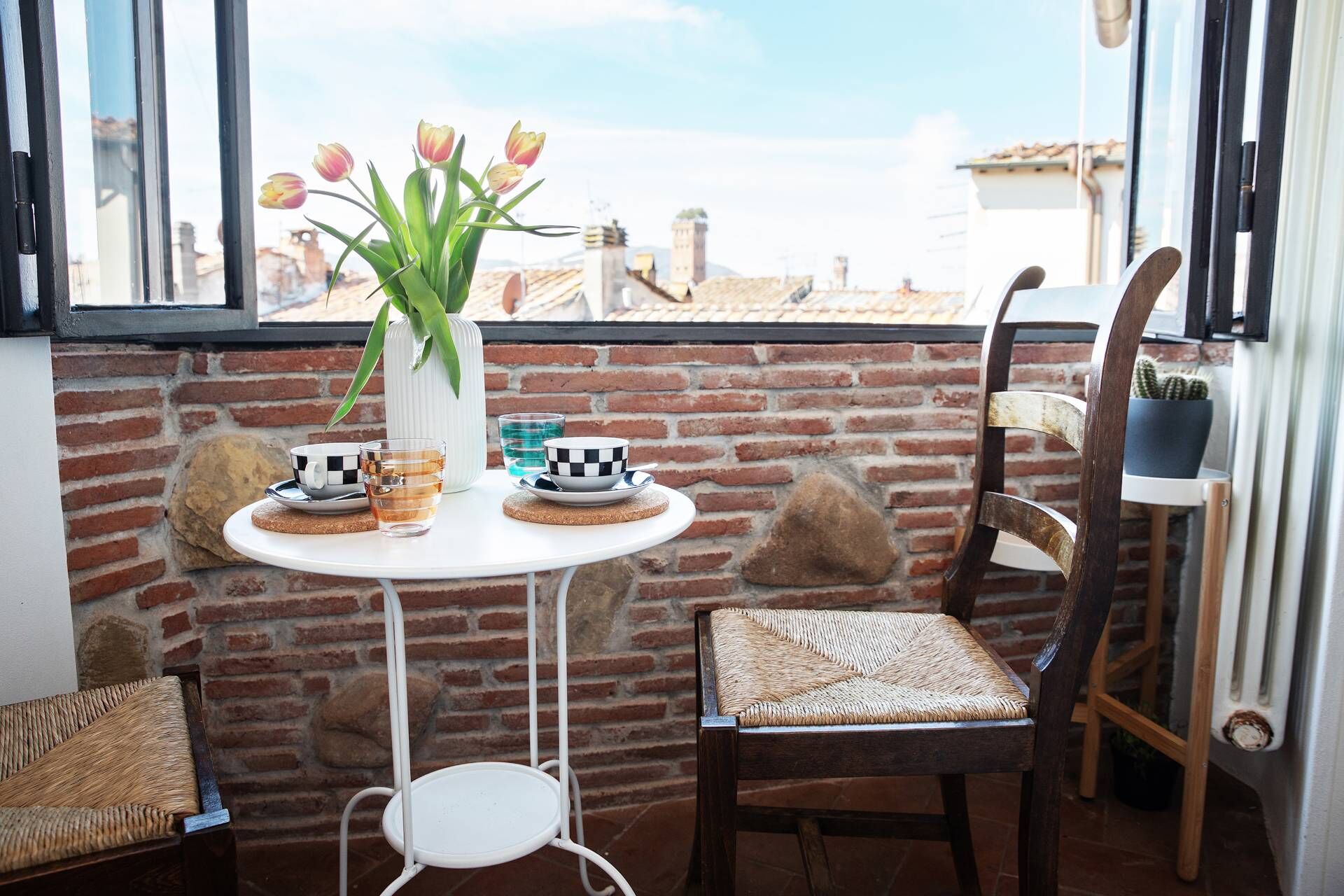 Un piccolo balcone con un tavolo bianco, due sedie, un vaso con tulipani e vista sugli edifici.