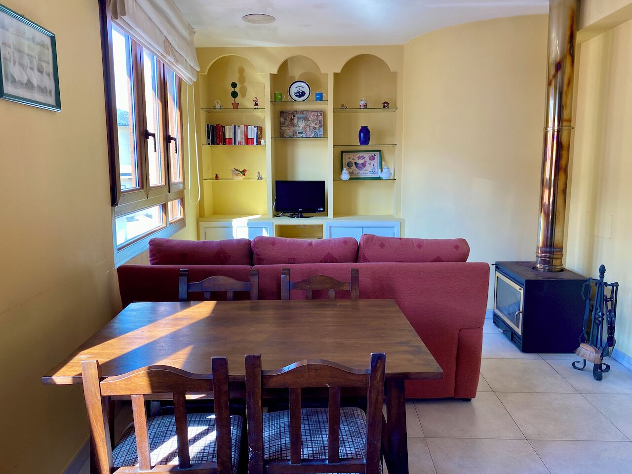 A dining area with a wooden table and chairs, a red sofa, and a television on a shelf.