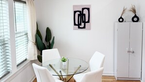 Een eetgedeelte met een ronde glazen tafel, witte stoelen, een wit kastje en een abstract kunstwerk aan de muur.