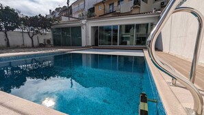 Una piscina con una escalera metálica, rodeada por un deck de baldosas y un edificio con ventanas amplias.