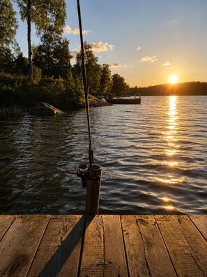 A wooden dock with a fishing rod and reel, a calm body of water, and a sunset in the background. - A Stunning Swedish House by the Lake • Fireplace, Boat & Separate Guest House (Belganet)
