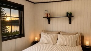 Un dormitorio con cama de madera, ventana con vista a un lago, estante en la pared con objetos decorativos y un respiradero en el techo.