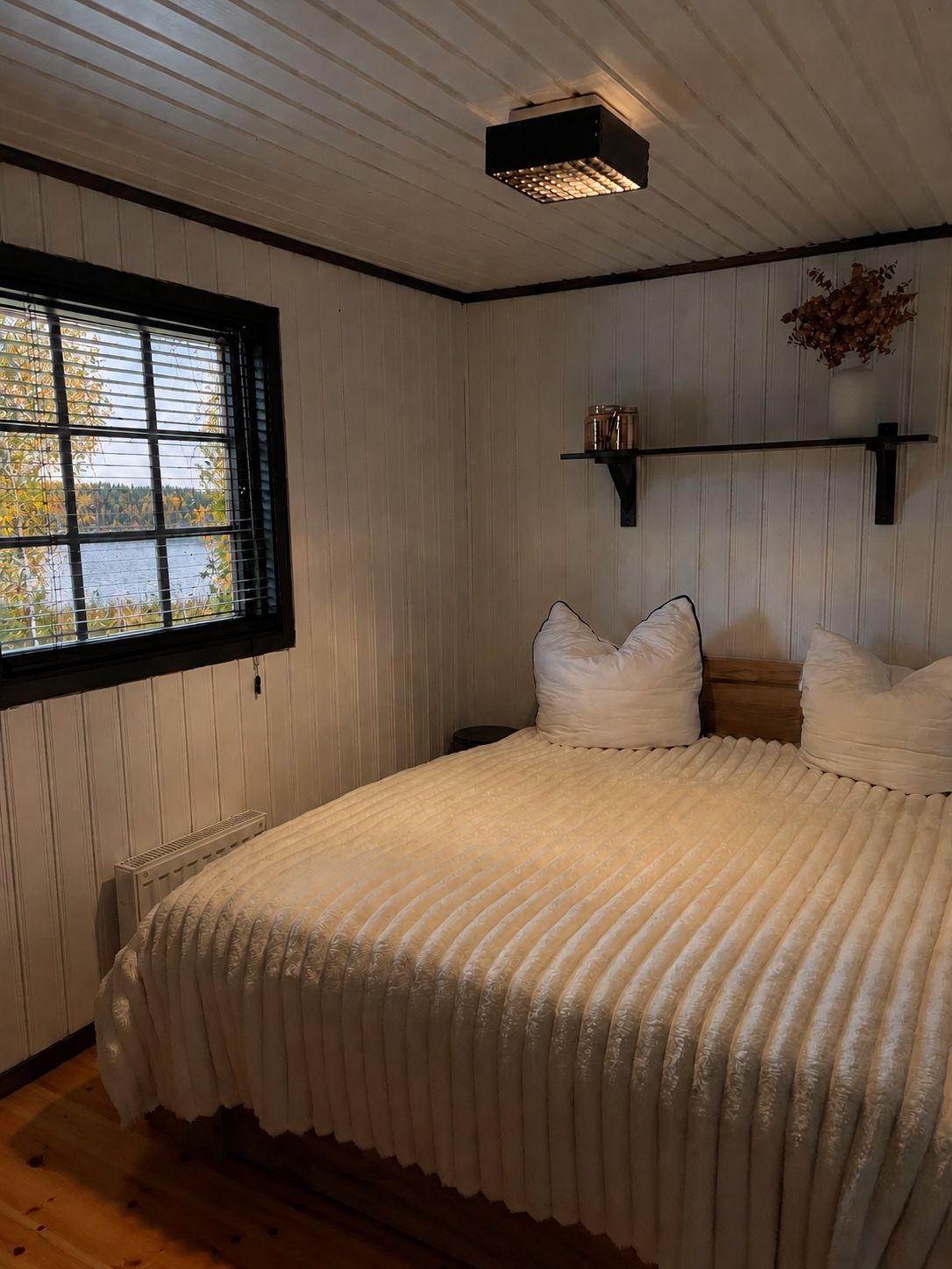 A bedroom with a bed, a window with blinds, a shelf with jars, and a ceiling light.
