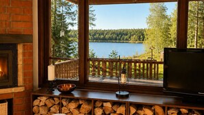 Una habitación acogedora con chimenea, un ventanal grande con vistas a un lago, televisión y una pila de leña.