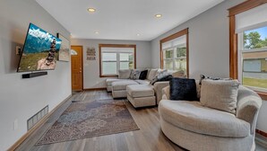 A spacious living room with a sectional sofa, a flat-screen TV mounted on the wall, and a large window providing natural light.