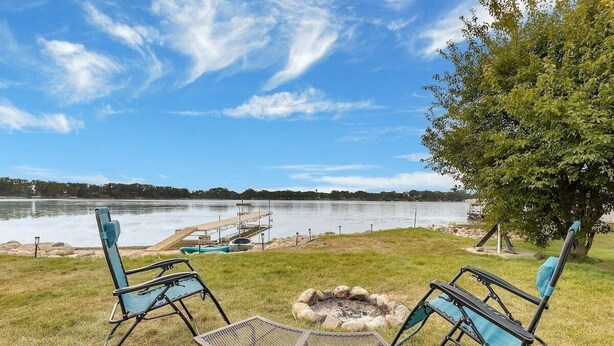 A lakeside outdoor seating area with two chairs and a table, a fire pit, and a dock extending into the water.