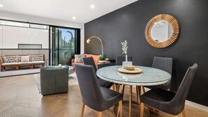 Een moderne eetruimte met een ronde glazen tafel, grijs gestoffeerde stoelen en een zwarte accentmuur met een spiegel met houten lijst.