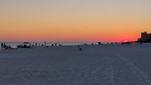 En strand med folk och parasoller, byggnader i bakgrunden och en solnedgång.