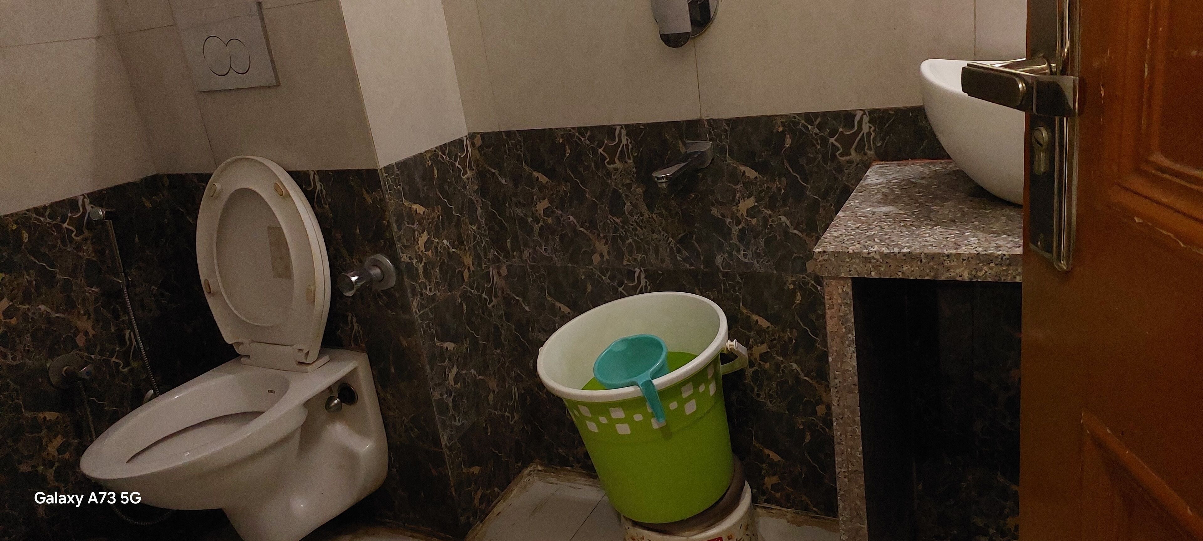 Une salle de bain avec des toilettes, un seau vert et un mur en marbre.