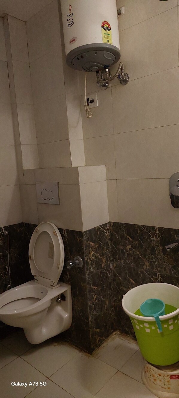 Une salle de bain avec des toilettes, un seau vert et un chauffe-eau fixé au mur.
