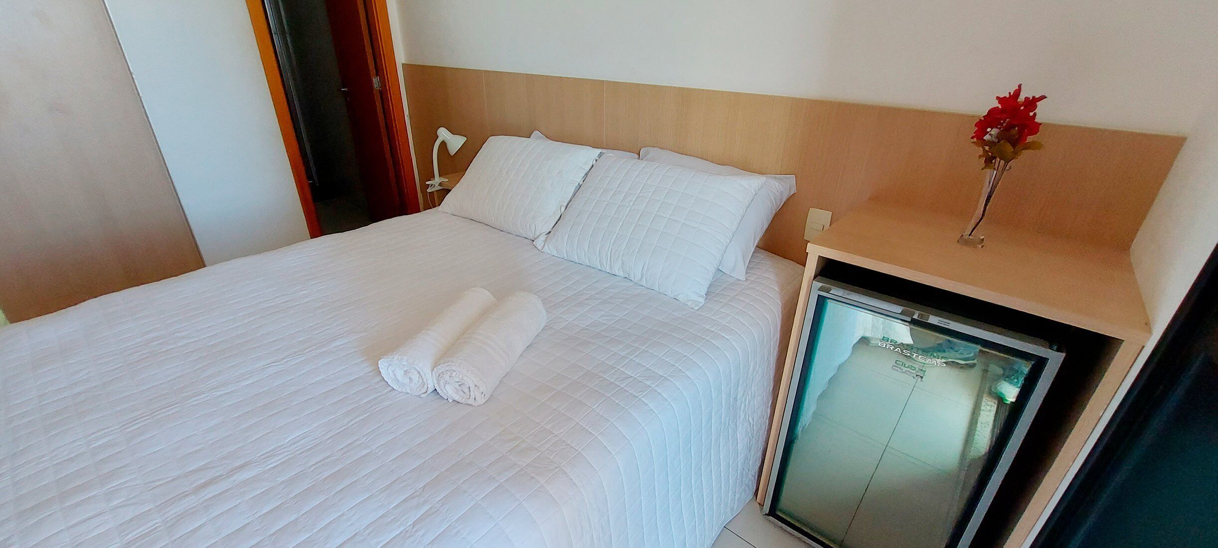 A neatly made bed with white linens and pillows, a bedside table with a vase of flowers, and a small refrigerator.
