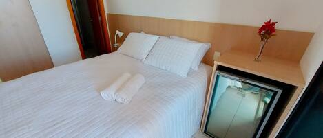 A neatly made bed with white linens and pillows, a bedside table with a vase of flowers, and a small refrigerator.