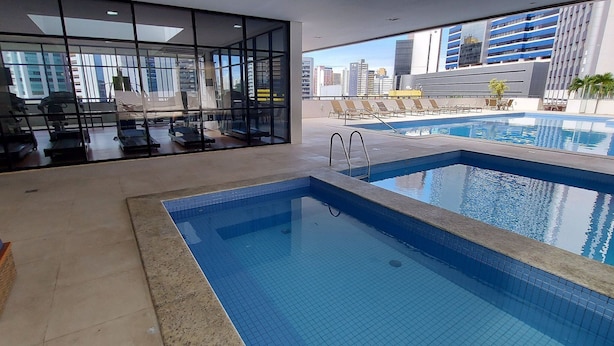 Un espace piscine intérieur moderne, doté d’équipements de musculation, de chaises longues et offrant une vue sur le paysage urbain.