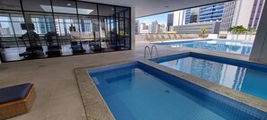 Un área interior de piscina moderna con equipo de ejercicio, sillones de descanso y vistas a la ciudad.