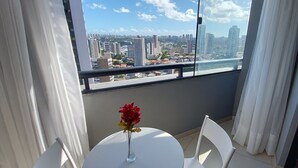 Un balcon avec une table et des chaises, un vase rempli de fleurs et une vue sur la ville par la fenêtre.