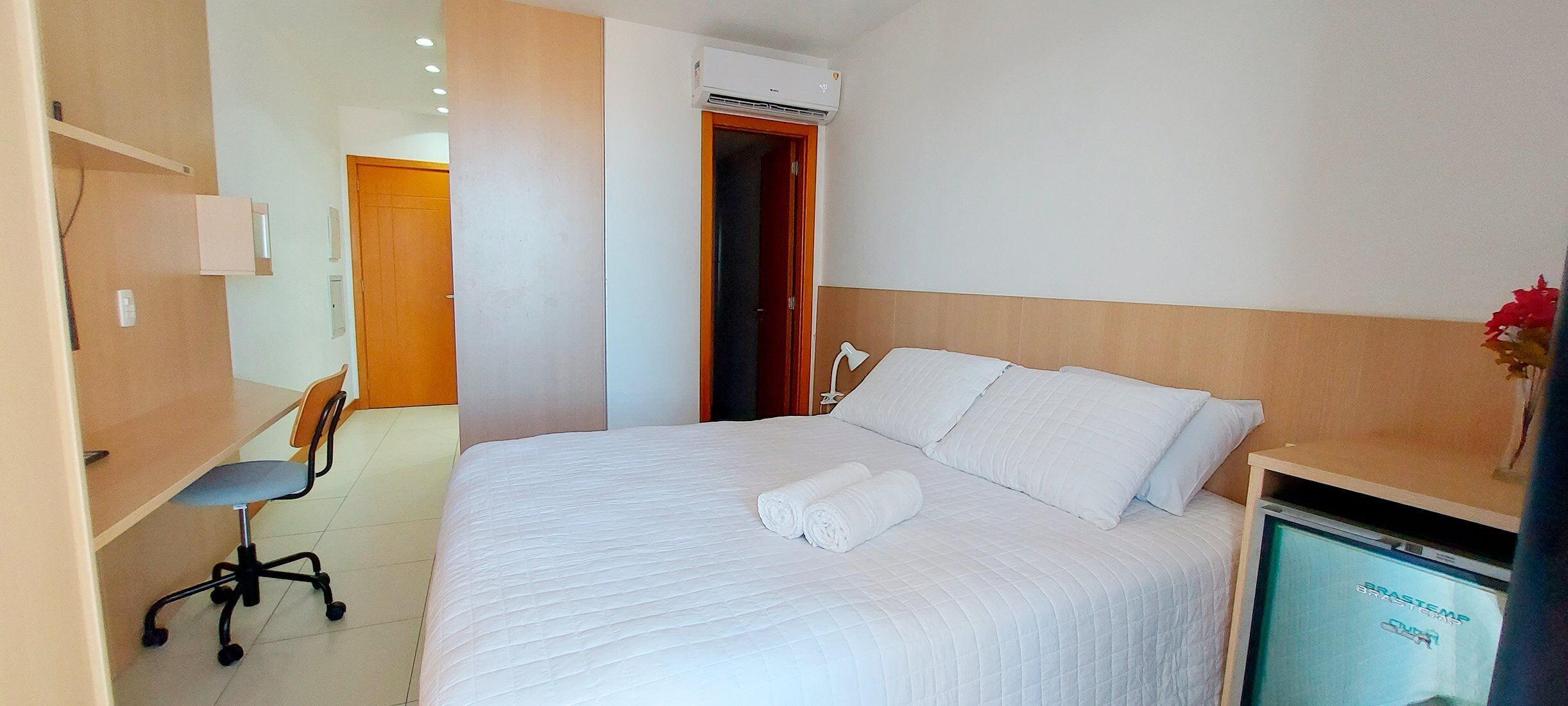 A hotel room with a bed, a desk, a chair, an air conditioning unit, and a television.