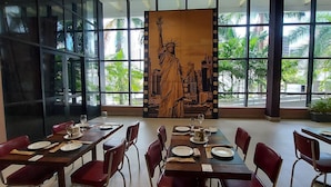 Un restaurant avec des tables dressées pour le dîner, de grandes fenêtres offrant une vue sur le paysage urbain et une fresque représentant la Statue de la Liberté.