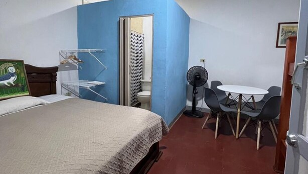 Una habitación pequeña y luminosa con una cama, una mesa con sillas, un ventilador y un baño con ducha.
