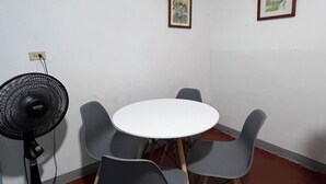 Una sala pequeña para reuniones con una mesa redonda y seis sillas, un ventilador de pie y dos cuadros en la pared.
