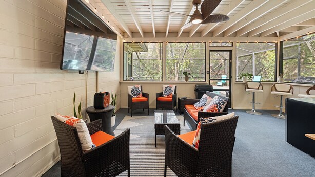 A sunlit patio with wicker furniture, a ceiling fan, and large windows offering a view of greenery. - Midcentury Marvel 5-bedroom pool house – walk to UF Stadium & Campus (Gainesville)