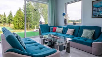 Un moderno salón con un sofá azul verdoso, una mesa de centro de vidrio y vistas a un césped verde y árboles a través de grandes ventanales.