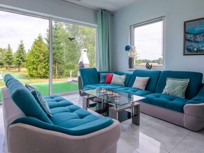 A modern living room with a teal sofa, a glass coffee table, and a view of a green lawn and trees through large windows. - Luxurious Holiday Home With Swimming Pool (gmina Dobra)
