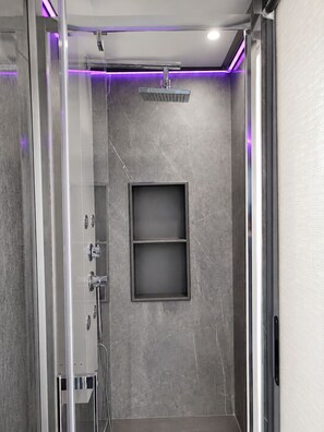 A modern shower with a glass enclosure, a wall-mounted shelf, and a ceiling light.