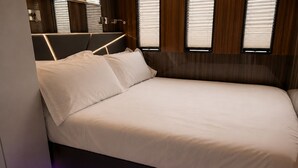 En moderne seng med sengegavl, et vindue med rullegardiner, og en lilla LED-lyslist langs gulvet.