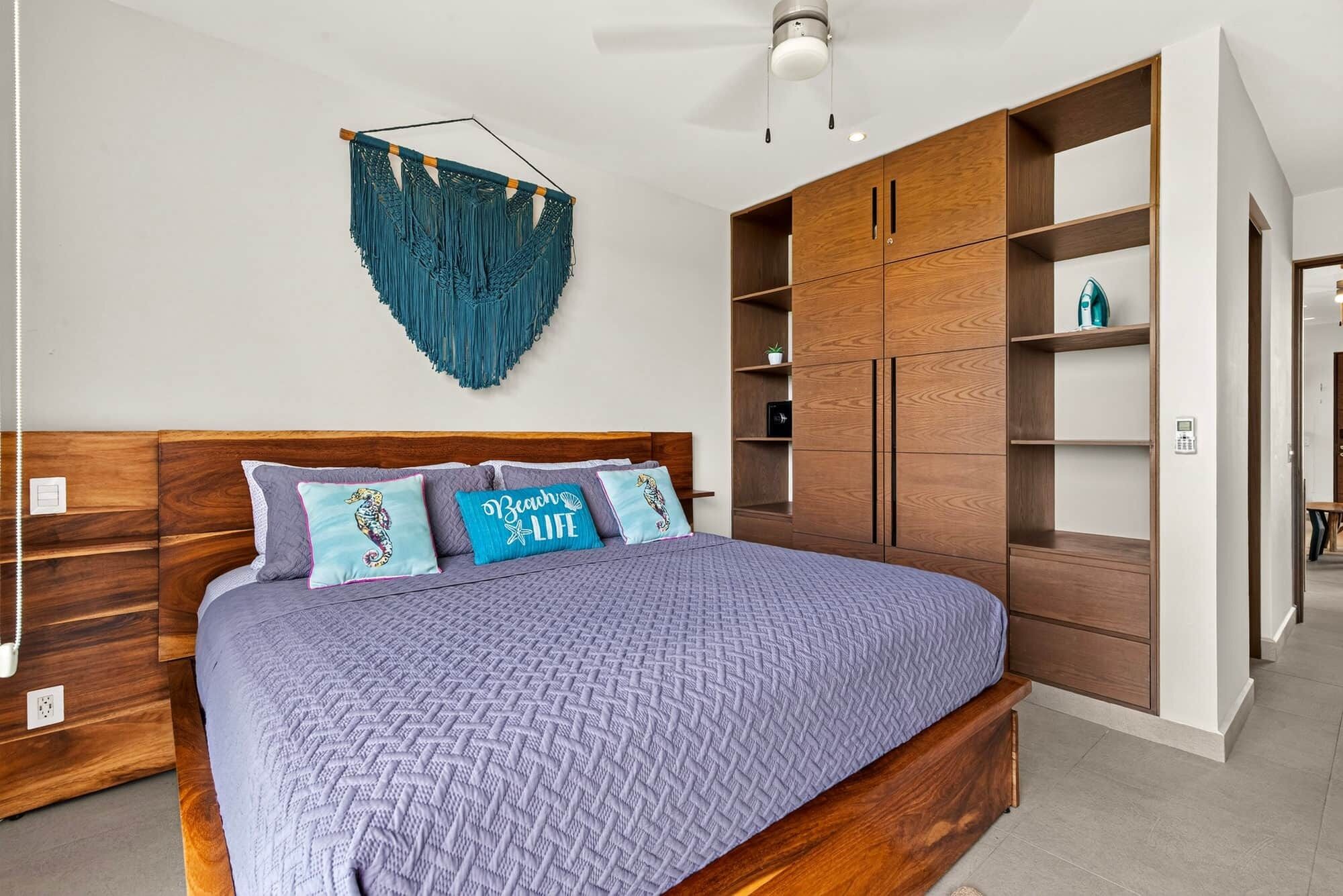 A bedroom with a wooden headboard, a bed with a patterned bedspread, and a wall-mounted macrame piece.