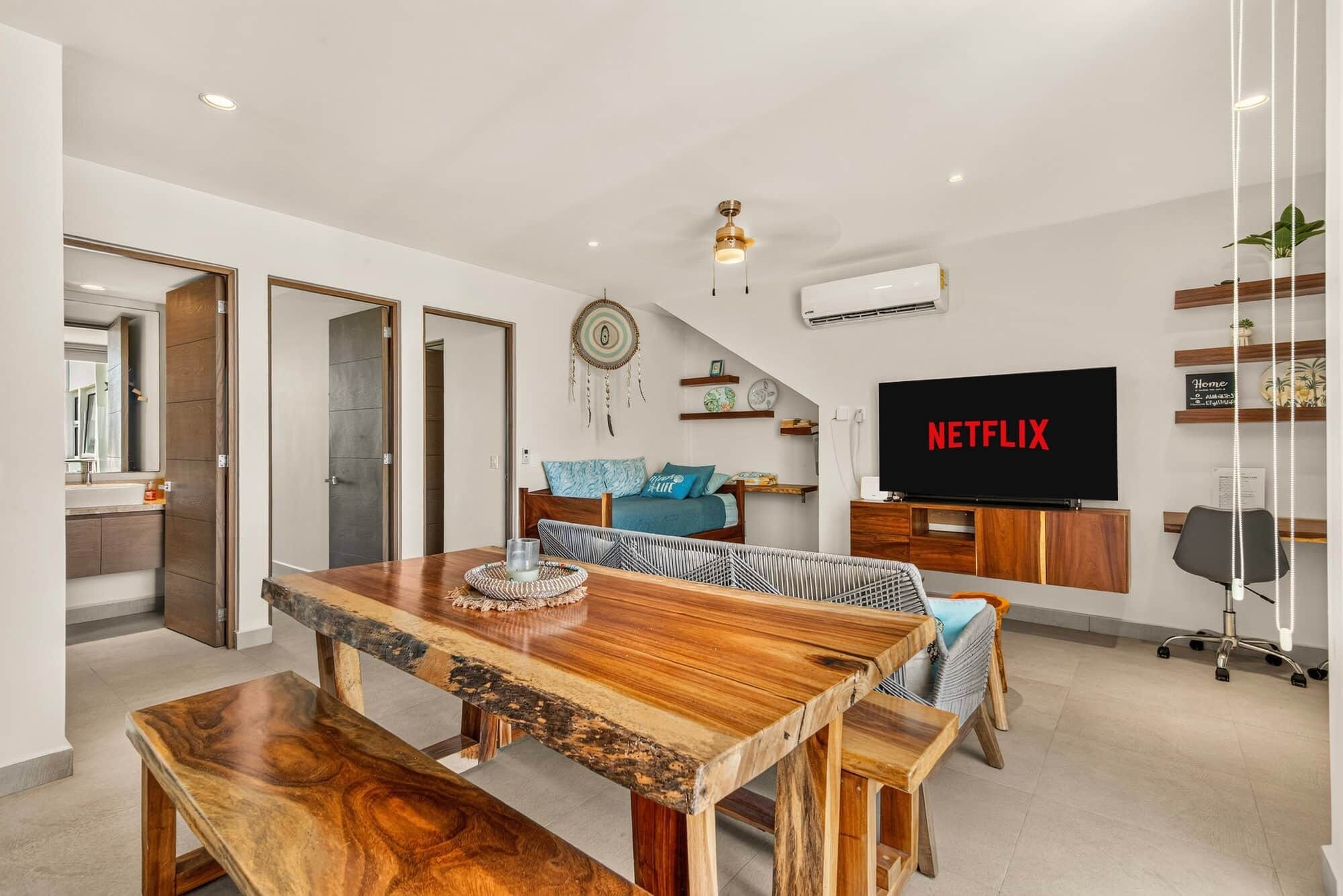 A modern living room with a wooden dining table, a sofa, a TV displaying 'NETFLIX', and a ceiling fan.
