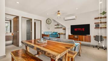 A modern living room with a wooden dining table, a sofa, a TV displaying 'NETFLIX', and a ceiling fan.