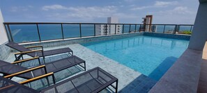 Ein Infinity-Pool auf dem Dach mit Glasgeländern, Liegestühlen und Blick auf das Meer und die Skyline.