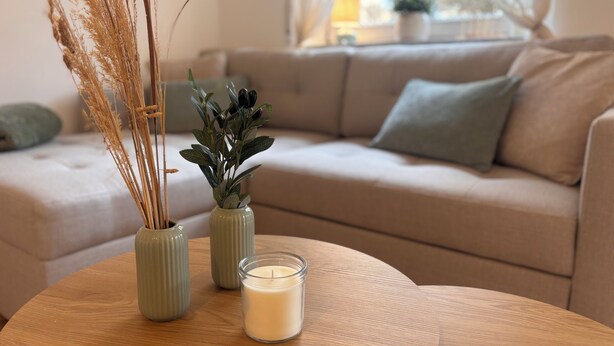 Ett vardagsrum med en beige soffa, ett runt träbord och två gröna vaser med torkade växter och ett ljus.