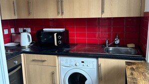 A compact kitchen with wooden cabinets, a washing machine, a toaster, and a red tiled backsplash. - Room 6 in Camden Parkway (London)