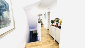 A narrow hallway with a wooden floor, white walls, and a staircase leading to an upper level. There is a framed picture on the left and a cabinet with plants on the right. - Room 6 in Camden Parkway (London)