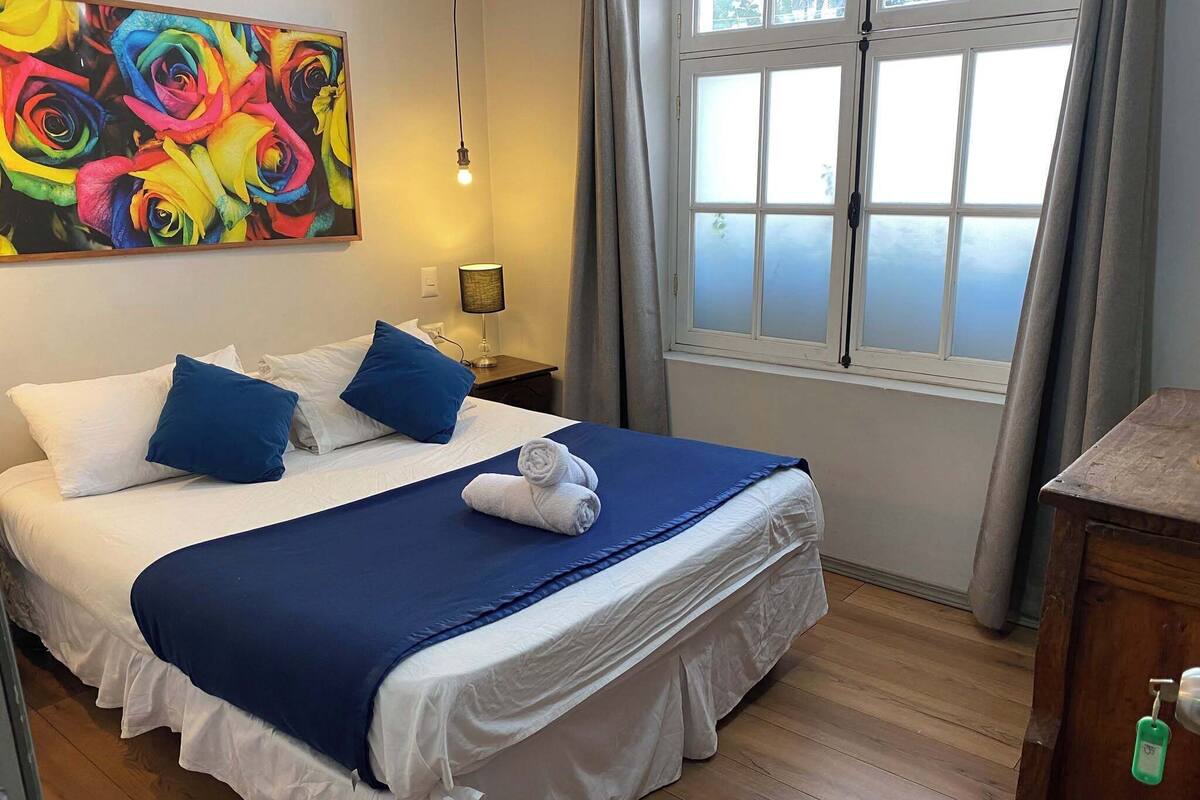 Een hotelkamer met een bed, kussens, een schilderij, een lamp, een raam en een houten vloer.